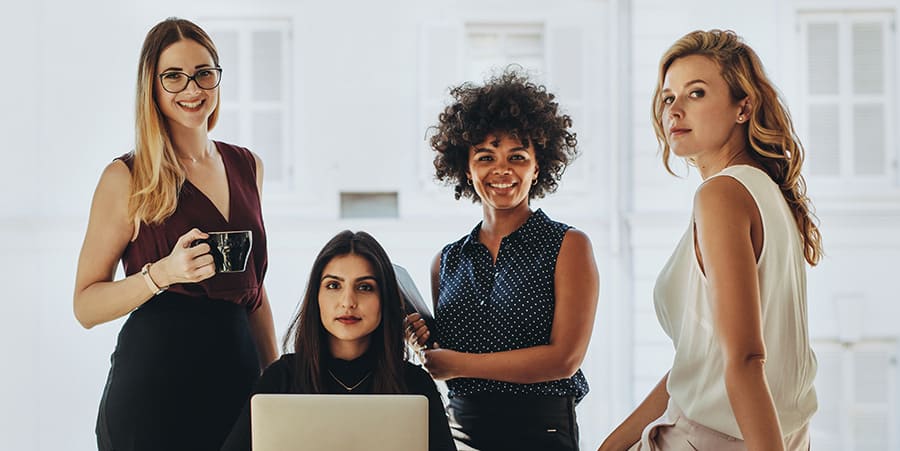 Four girls look confidently into the room, smiling slightly. They all sit around a desk, on which a laptop, coffee cups and office supplies are placed.