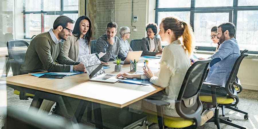 A diverse group of people sit around a table and engage in conversation. On the table are laptops, notebooks and pens. They are in an office environment and are dressed appropriately. 